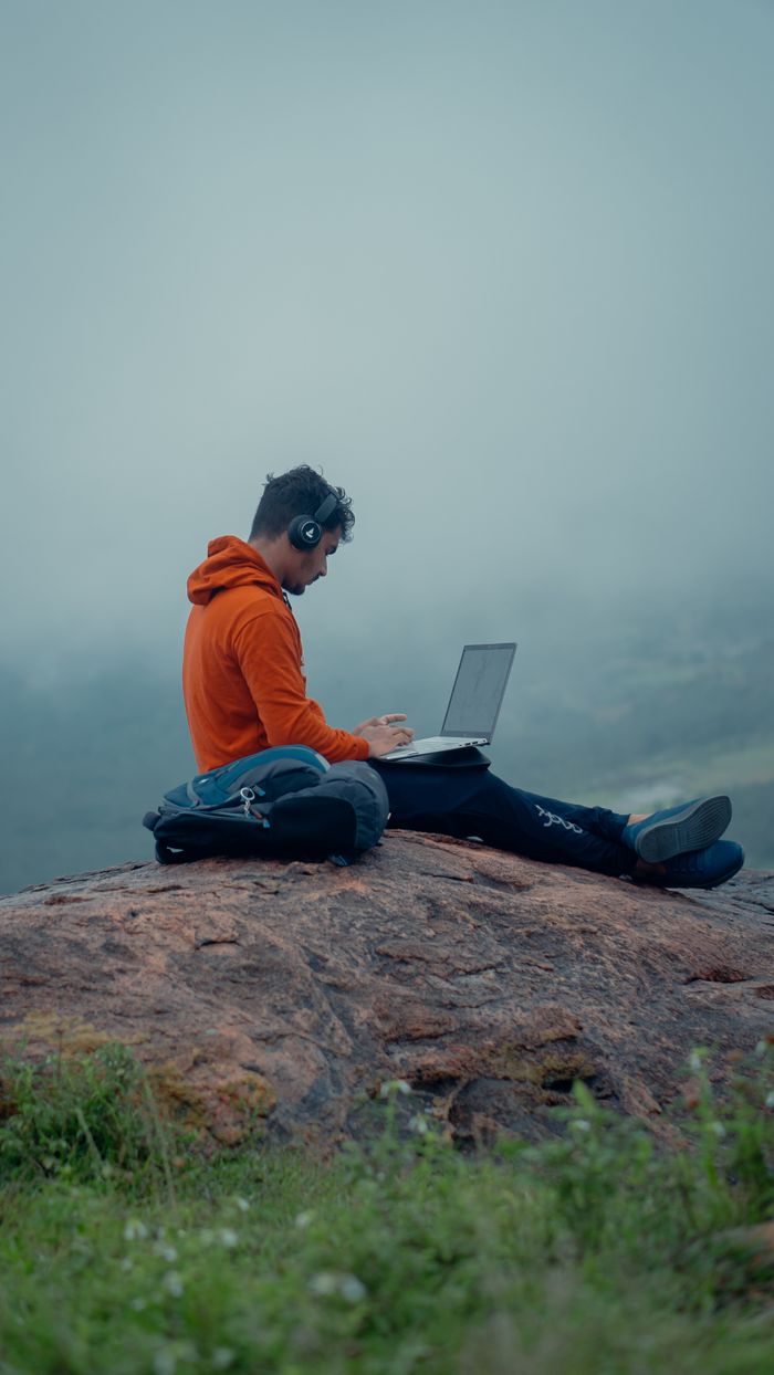 Man seated on a rock with laptop and headphones, embracing remote work in nature.